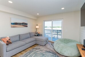 Una sala de estar moderna con un sofá seccional gris, un sillón de judío, un cuadro de un muelle, una lámpara de pie y un balcón con vistas.