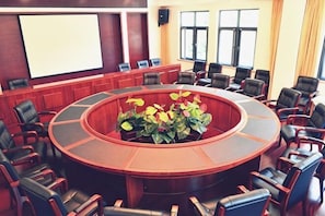 Ein Konferenzraum mit einem Tisch in der Mitte, umliegenden Stühlen, einer Projektionsleinwand und Fenstern.