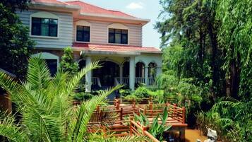 A two-story house with a red roof, surrounded by lush greenery and a wooden bridge.