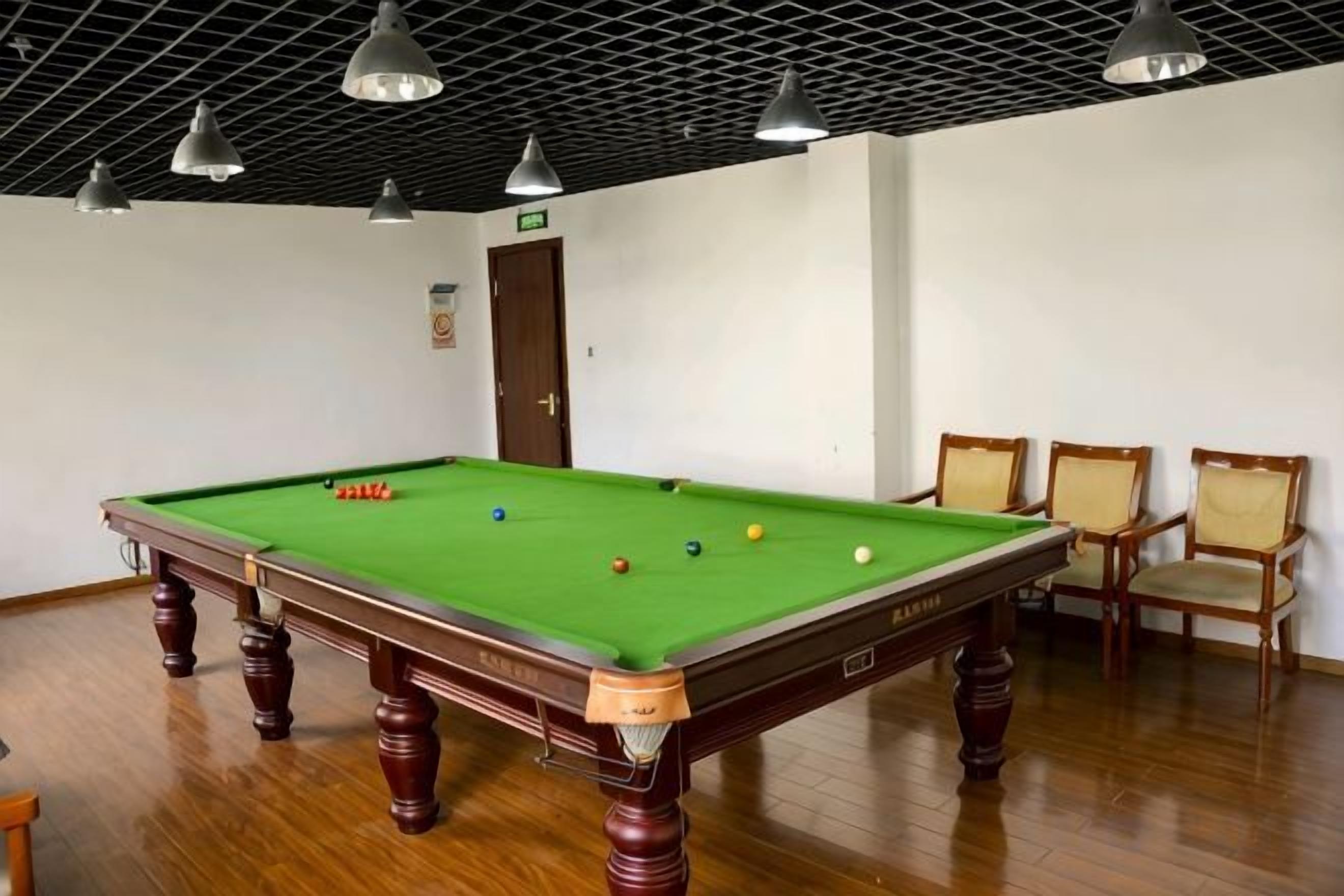 A pool table with balls, chairs, and a door in a room with wooden flooring and ceiling lights.