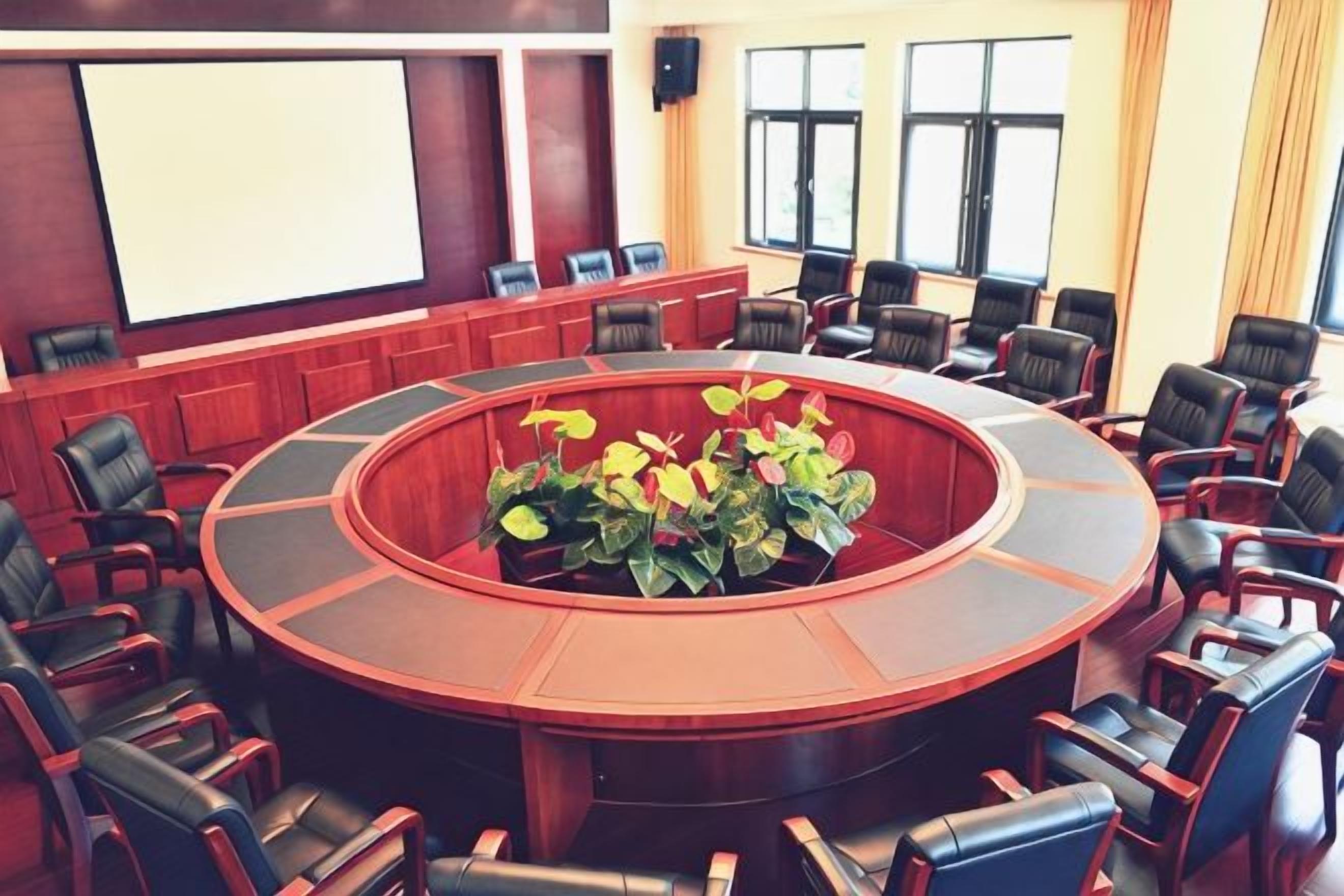 Un salón de conferencias con una mesa circular rodeada de sillas, una pantalla de proyector y un ventanal con cortinas.