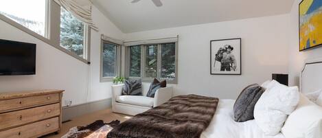 A bedroom with a bed, a ceiling fan, a TV, a window, and a painting.