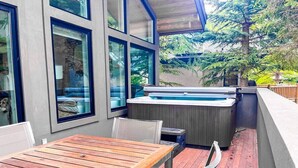 Une terrasse extérieure avec une table et des chaises en bois, un jacuzzi et une vue sur les arbres et une maison.