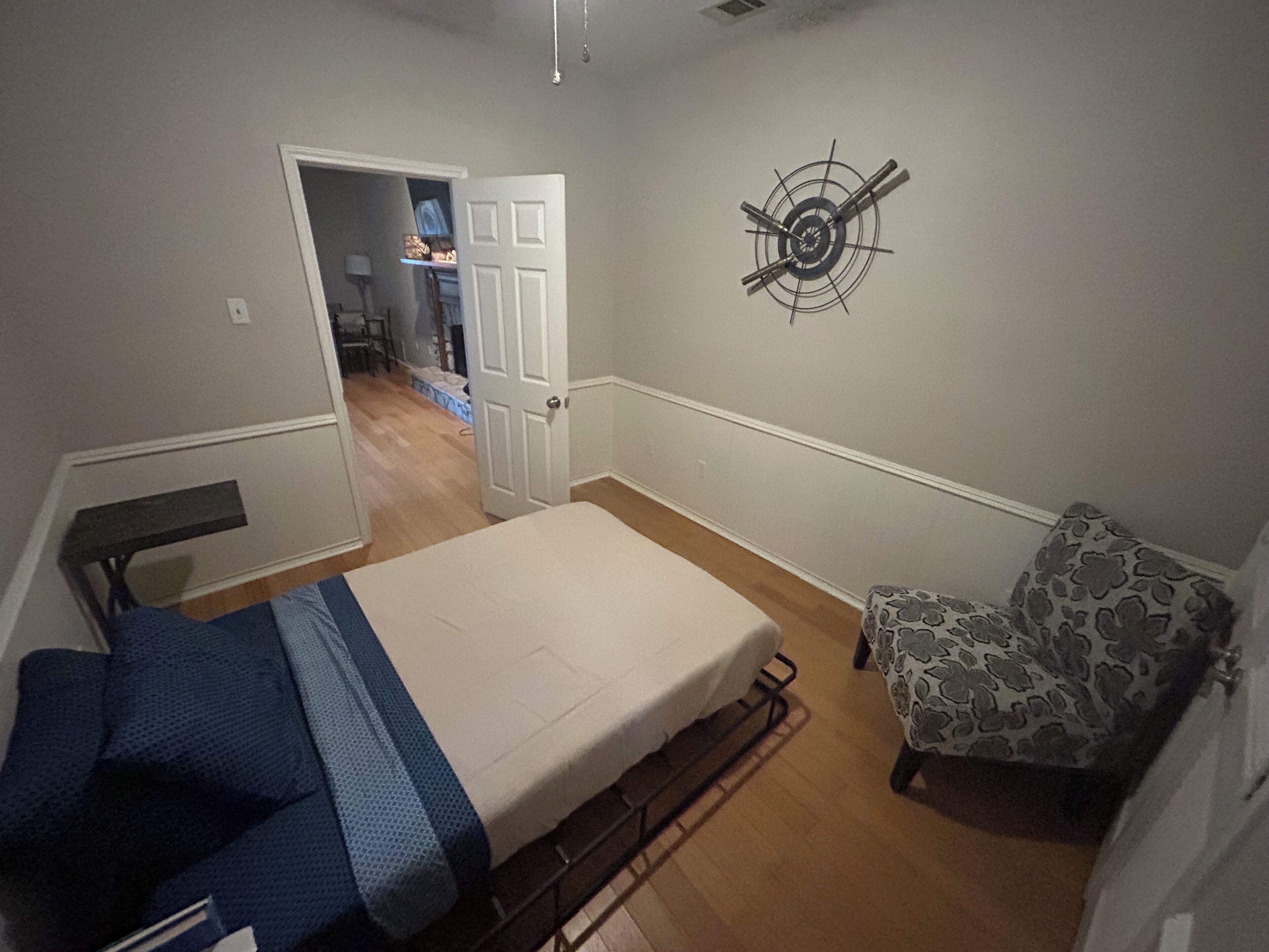 Pequeño dormitorio con cama, una silla, un reloj de pared y una puerta que da a otra habitación.