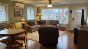 A living room with a sectional sofa, a dining table with chairs, a ceiling fan, and large windows with blinds. - Port Angeles History House (Port Angeles)