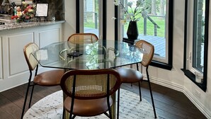 A modern kitchen with white cabinets, a glass dining table, wicker chairs, and a view of the outdoors through large windows. - Beautiful Deck with Golf Course View/ Game Room/ Fire Pit (Conroe)