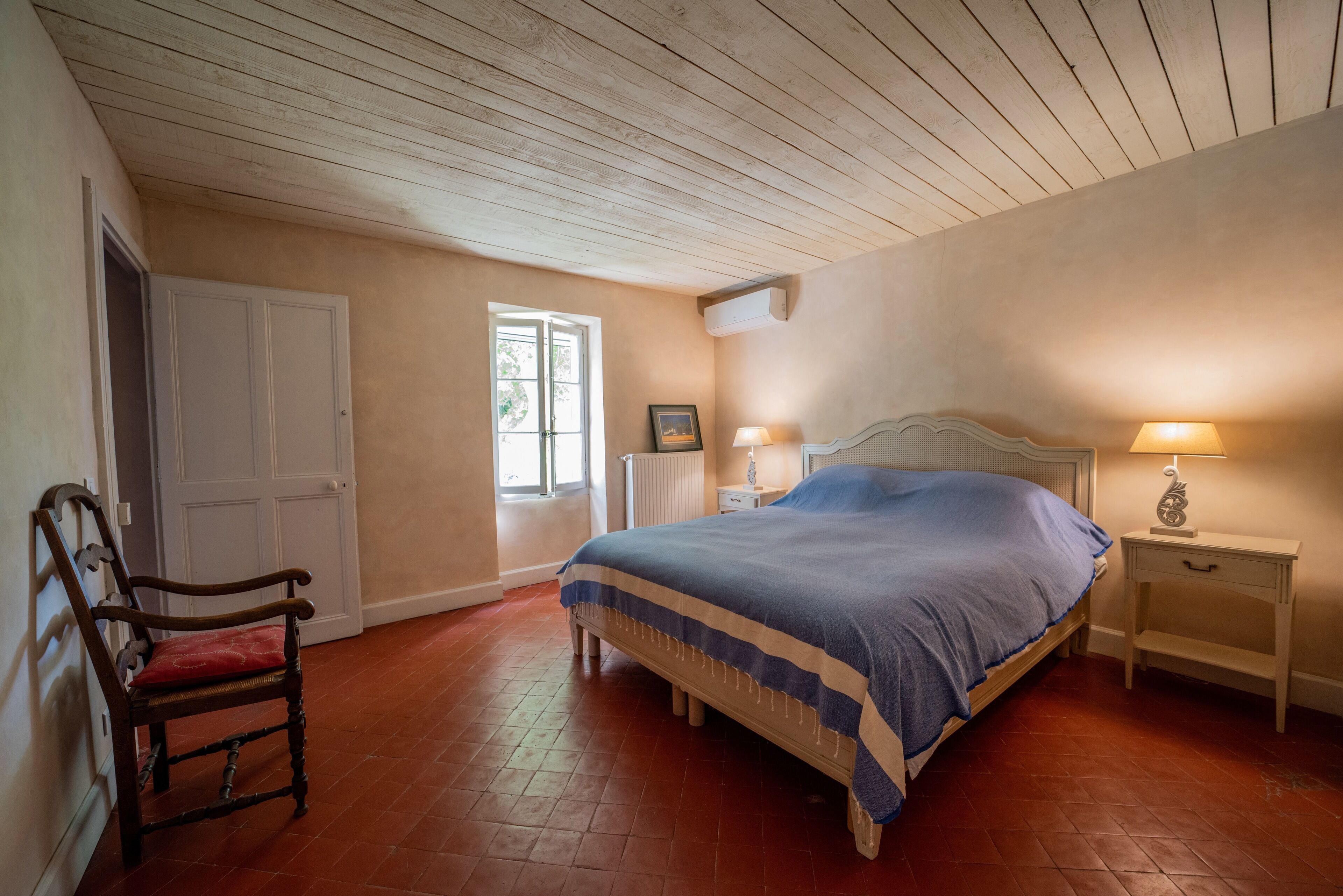 Un dormitorio con techo de madera, una cama con cubrecama azul, una silla, una mesita de noche con lámpara y una ventana con persianas.