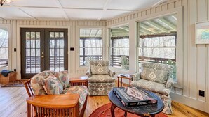 A cozy living room with a wooden floor, a dark door, and large windows with a view of trees. There are two armchairs, a sofa, and a coffee table with a board game. - Beautiful Highlands Cabin: Large Game Room, Hot Tub (Highlands)