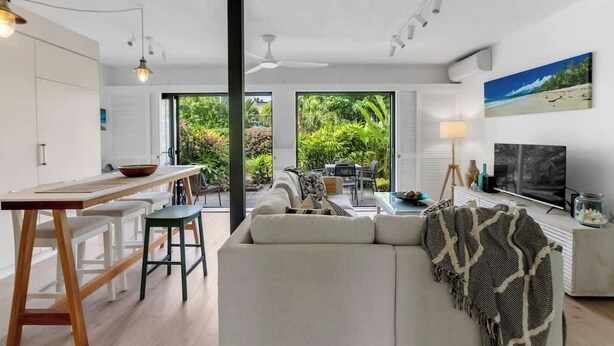 A modern living room with a sofa, dining table, and chairs, a TV, and a balcony with a view of greenery.