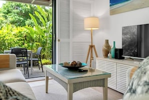 A modern living room with a glass-top table, a floor lamp, a television, and a view of lush greenery outside.