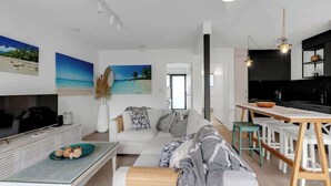 A modern living room with a sofa, coffee table, and a TV. There is a kitchen area with a dining table and chairs.