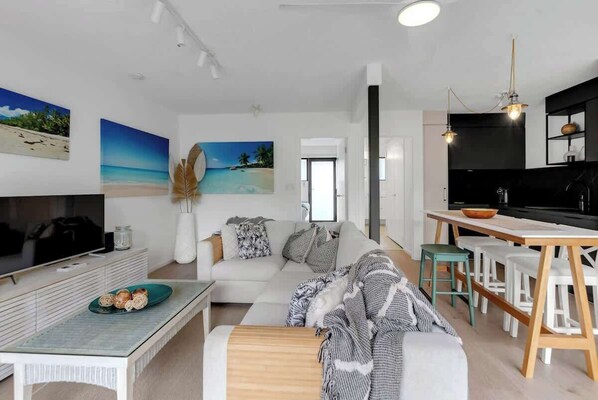 A modern living room with a sofa, coffee table, and a TV. There is a kitchen area with a dining table and chairs.