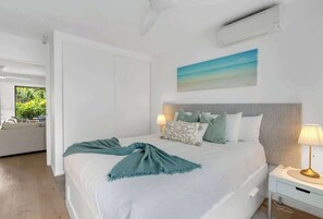 A modern bedroom with a large bed, a bedside table with a lamp, a painting on the wall, and a view of greenery outside.