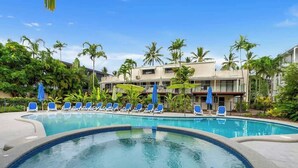 A swimming pool with blue lounge chairs, palm trees, and a building with balconies in the background.