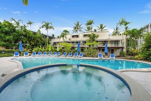 A swimming pool with blue lounge chairs, palm trees, and a building with balconies in the background.
