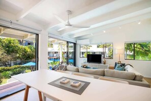 Een moderne woonkamer met een bank, een eettafel en een televisie. Er is een balkon met uitzicht op groen en een zwembad.