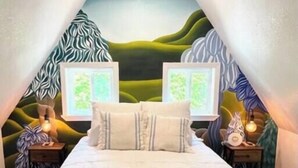 A bedroom with a sloped ceiling, a bed with a blue and white checkered bedspread, two bedside tables with lamps, and a mural on the walls. - Tree Top Oasis Near Waco (Mart)