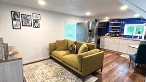 A modern living room with a yellow sofa, a patterned rug, and a kitchen with blue backsplash. - Tree Top Oasis Near Waco (Mart)
