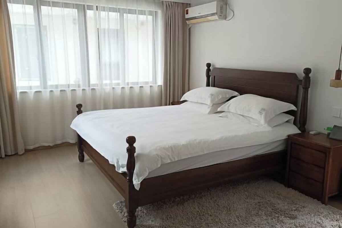 A bedroom with a wooden bed, white bedding, a nightstand, a lamp, and a window with curtains.