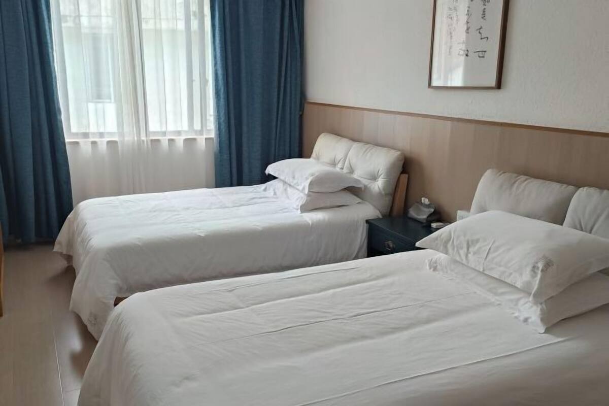 A hotel room with two beds, a nightstand, a framed picture on the wall, and a window with blue curtains.