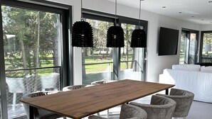 Un comedor moderno con una mesa de madera y sillas grises tapizadas, ventanas amplias que ofrecen vistas al exterior, y un sofá blanco al fondo.