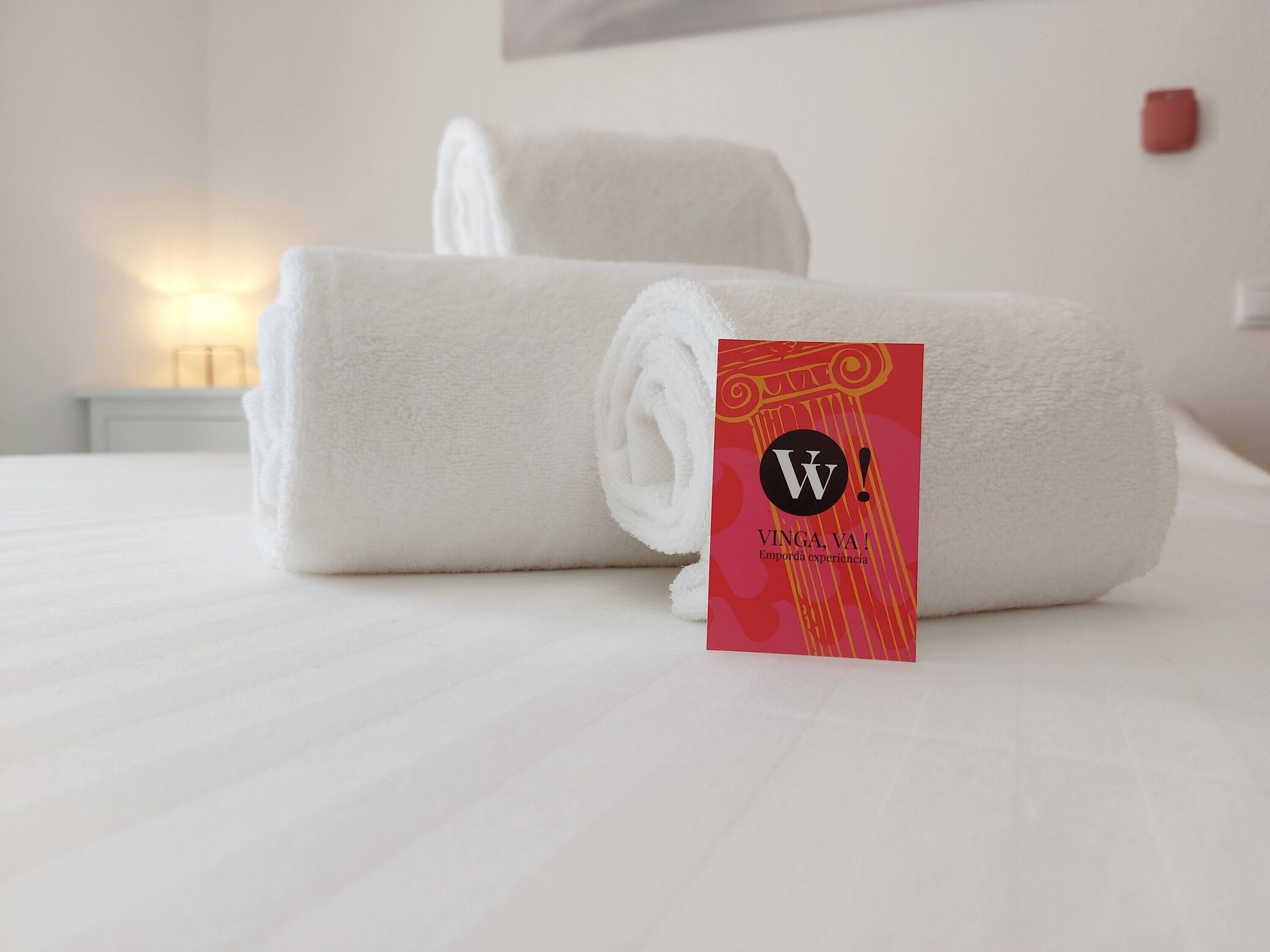 A neatly made bed with two rolled white towels and a red card with the text "VINGA, VA!" on a hotel room.