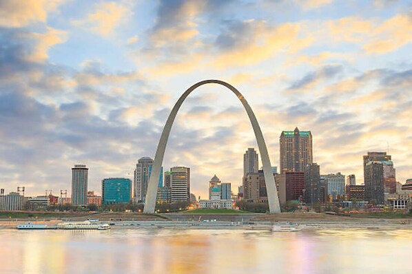 A city skyline with a prominent arch structure, modern buildings, and a calm river reflecting the sky.