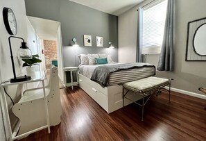 A bedroom with a bed, bedside tables, a desk, a chair, a plant, and artwork on the wall.