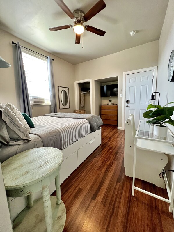 A bedroom with a bed, a desk, a ceiling fan, a TV, and a door.