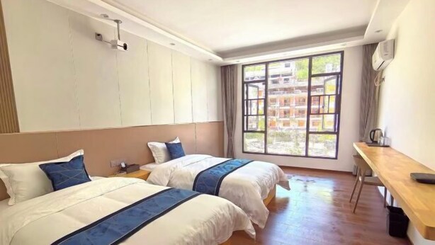 A hotel room with two beds, a wooden desk, a window with a view of buildings, and a ceiling-mounted air conditioning unit. - Lubianbian Inn (Zhangjiajie)
