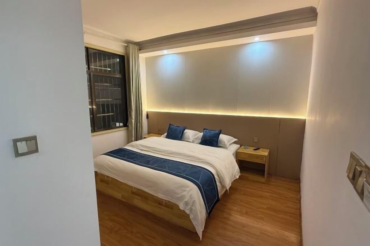 A bedroom with a bed, bedside tables, a nightstand, a window with curtains, and a wall-mounted light fixture.