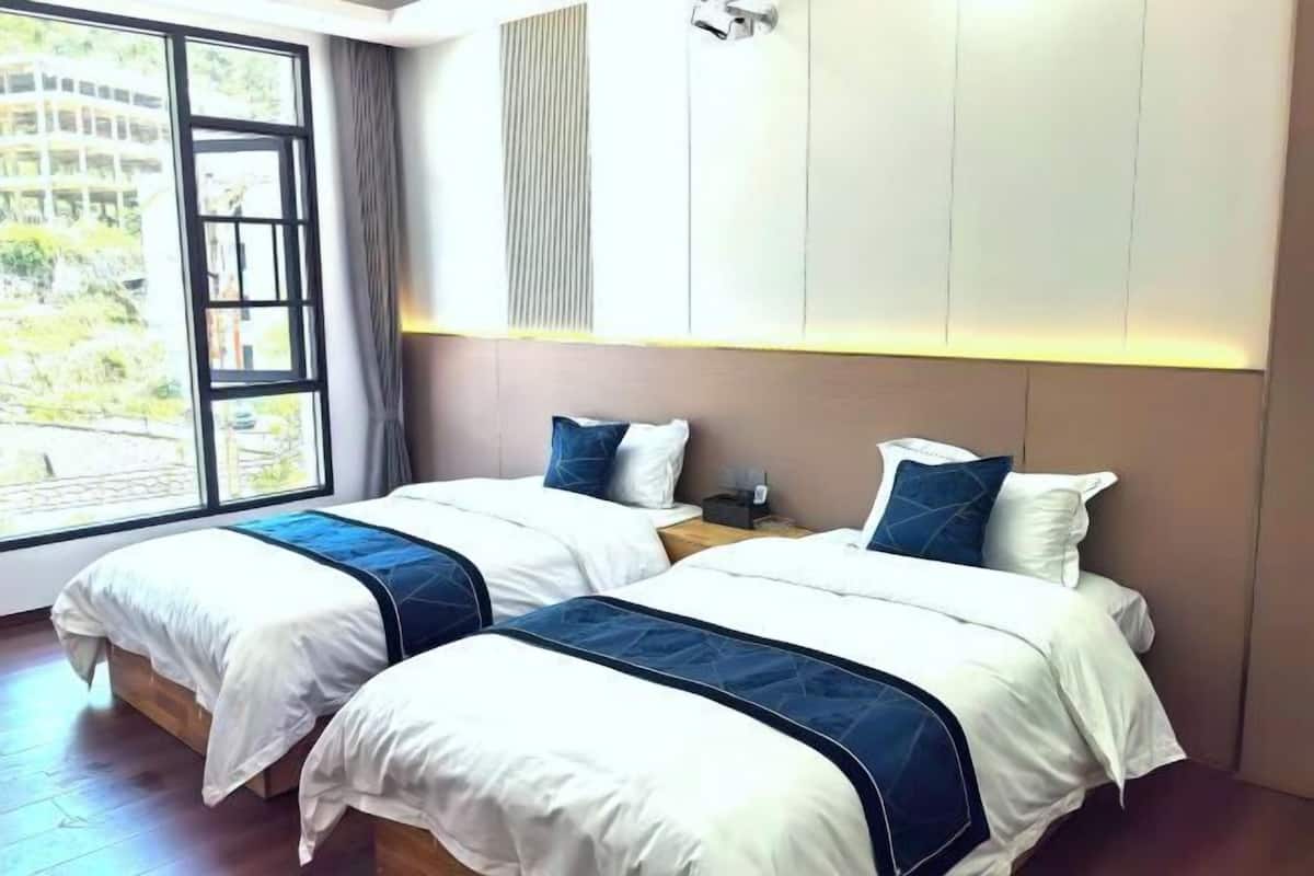 A hotel room with two beds, a large window, and a view of greenery outside.