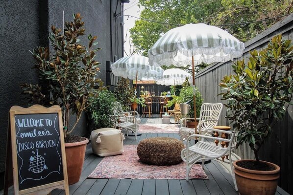 Une terrasse extérieure accueillante, avec des fauteuils en osier, des parasols à rayures et un panneau noir annonçant : « Bienvenue au jardin disco d’Emilee ».
