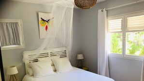 Un dormitorio con cama, ropa de cama blanca, una mesita de noche con lámpara, una obra de arte en la pared de un pájaro y una ventana con cortinas.