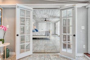 Quarto com cama, ventilador de teto e vista através de portas abertas para outro cômodo.