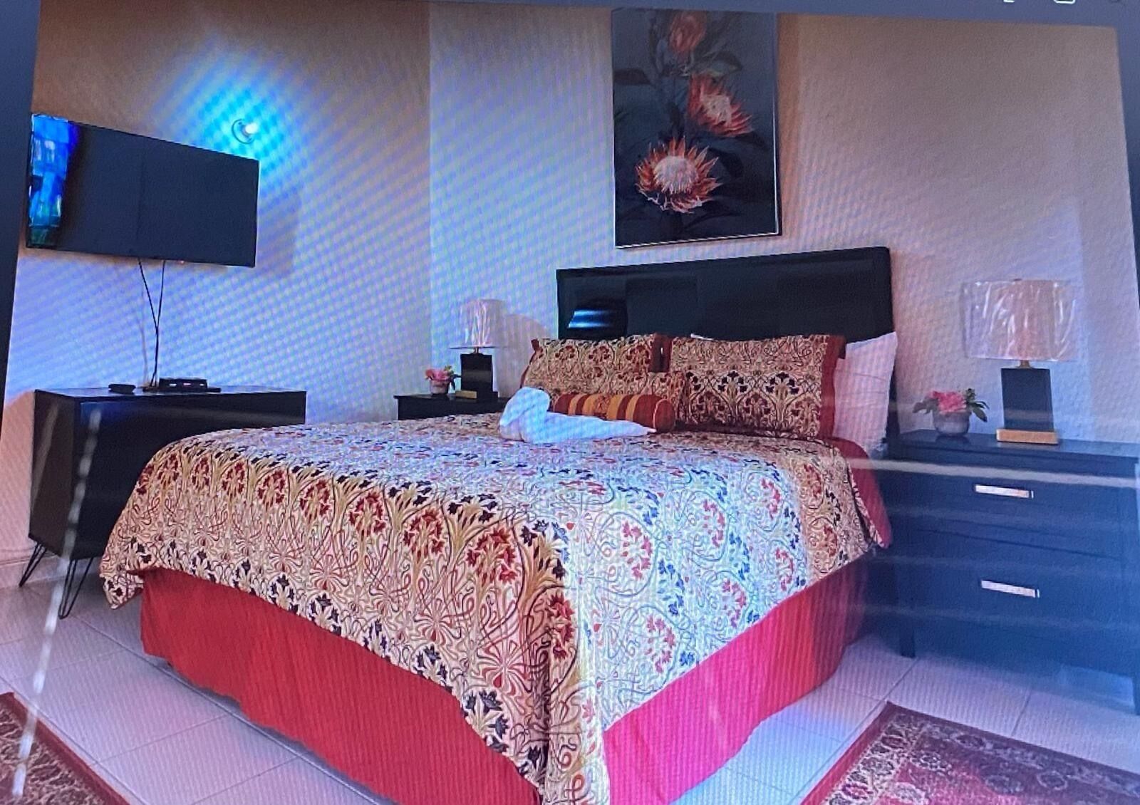 A bedroom with a large bed, a flat-screen TV mounted on the wall, and a nightstand with a lamp.