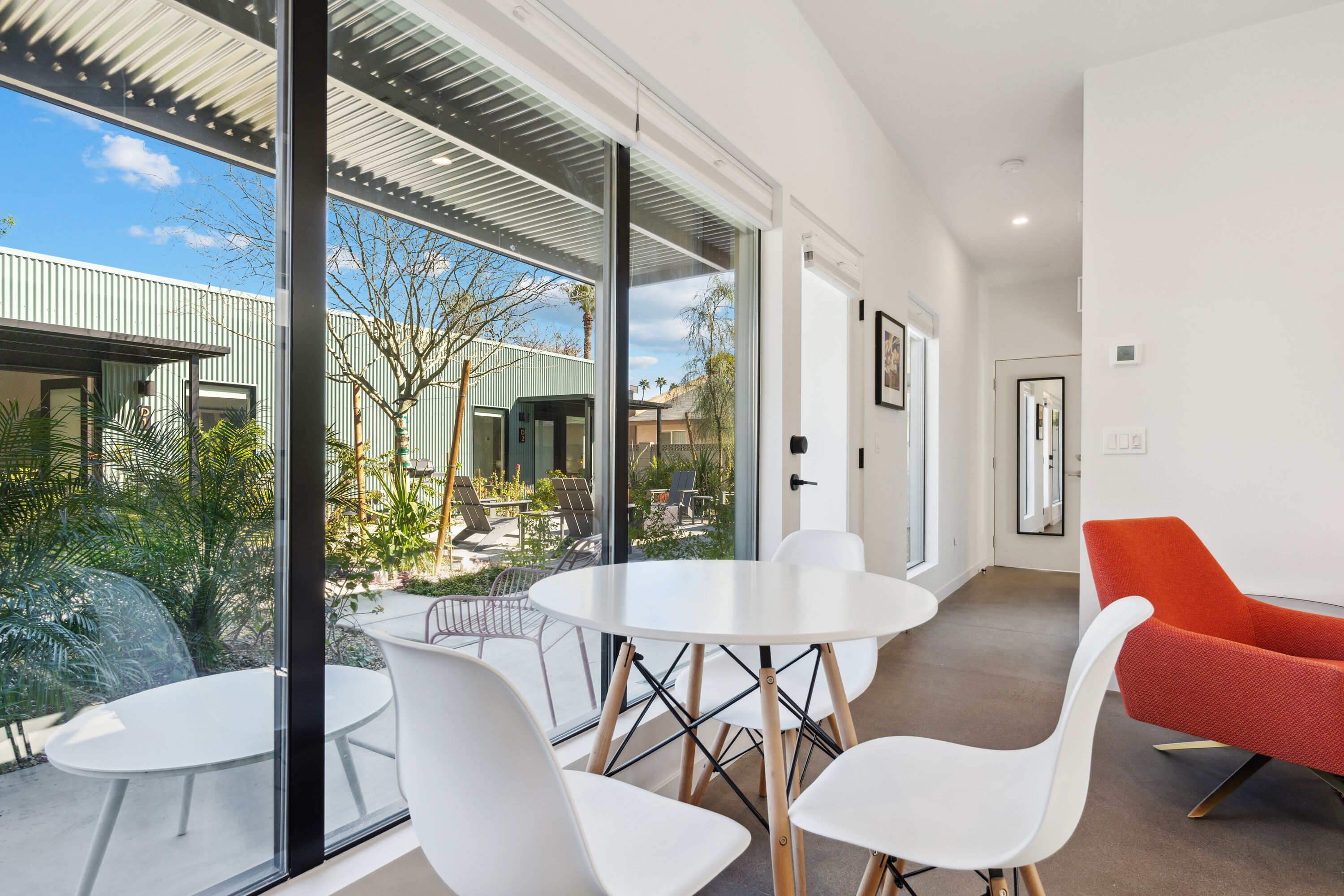 A modern interior with a round table and white chairs, large windows offering a view of a courtyard with plants and a building.