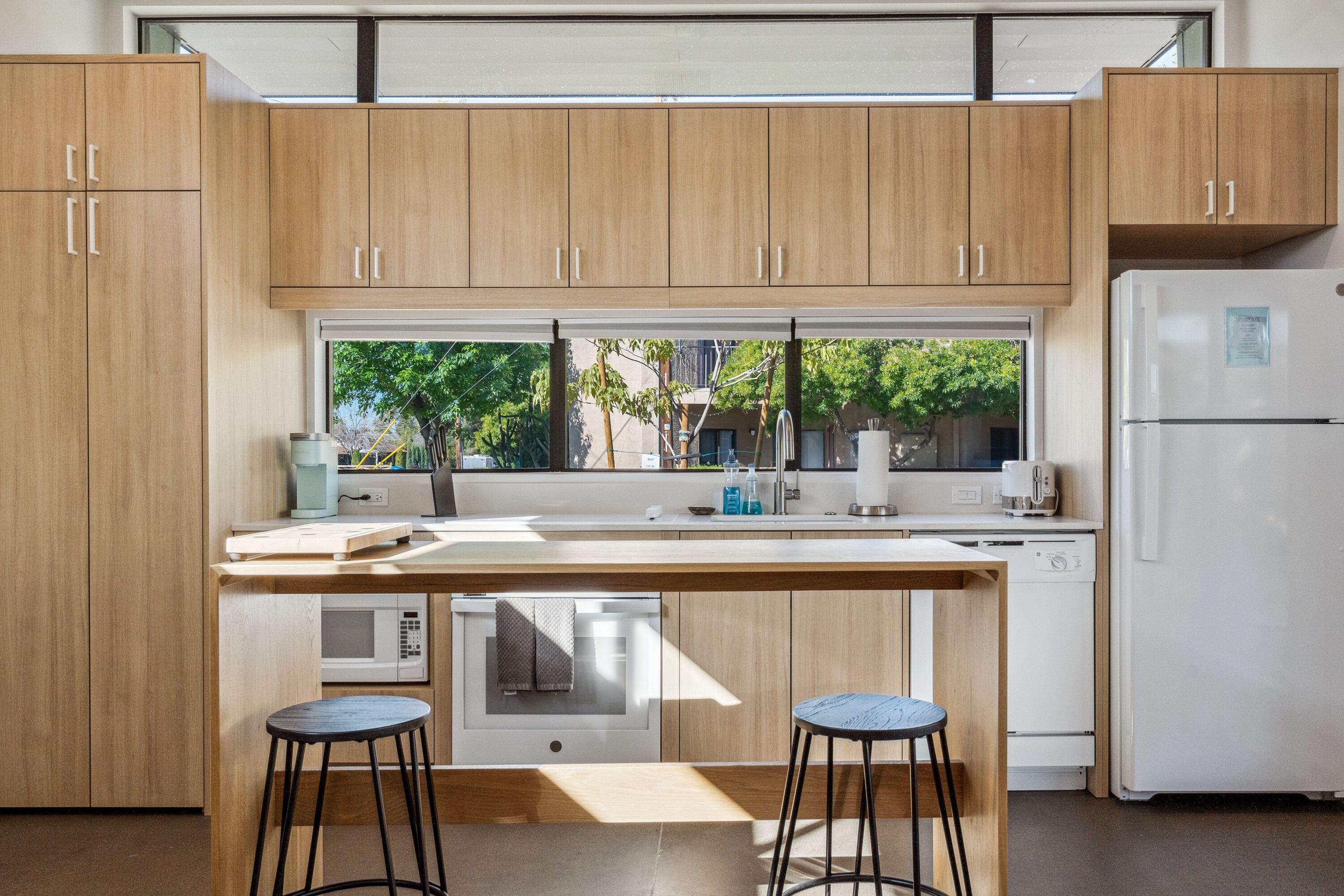 A modern kitchen with wooden cabinets, a central island with stools, a microwave, and a refrigerator.