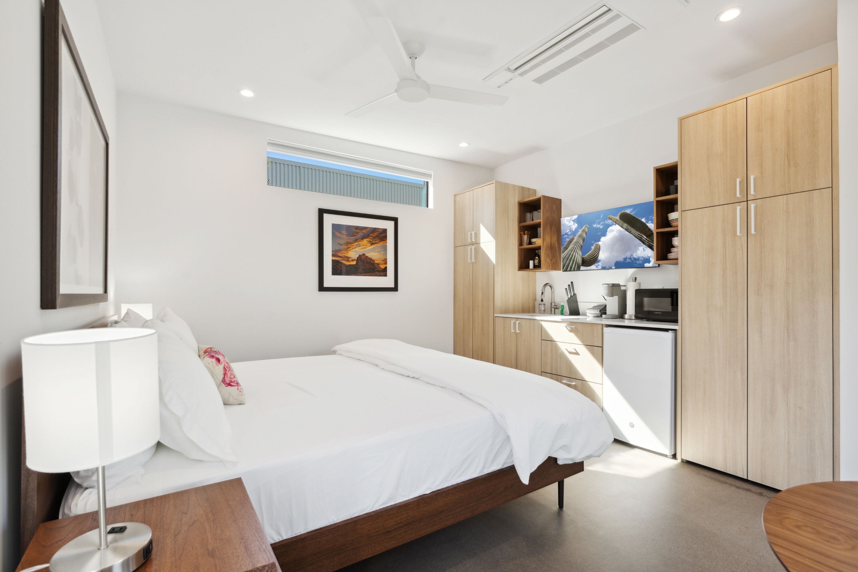 A modern bedroom with a bed, bedside table, lamp, and a view of a kitchen area with cabinets and a microwave.