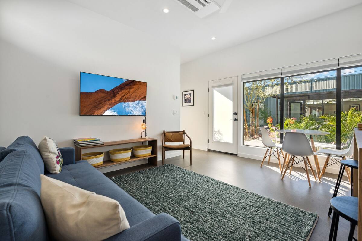 Una sala de estar moderna con sofá, una mesa de centro y un televisor montado en la pared. Hay una puerta corrediza de vidrio que da a un área exterior con una mesa y sillas.