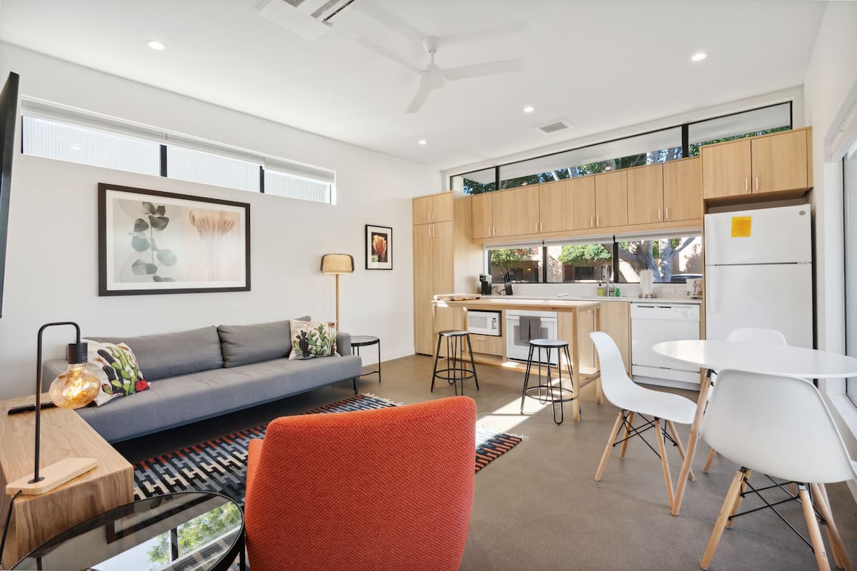 Una sala moderna con un sofá gris, una silla naranja y una pequeña mesa con una lámpara. Se observa una zona de cocina con armarios de madera y un refrigerador blanco.