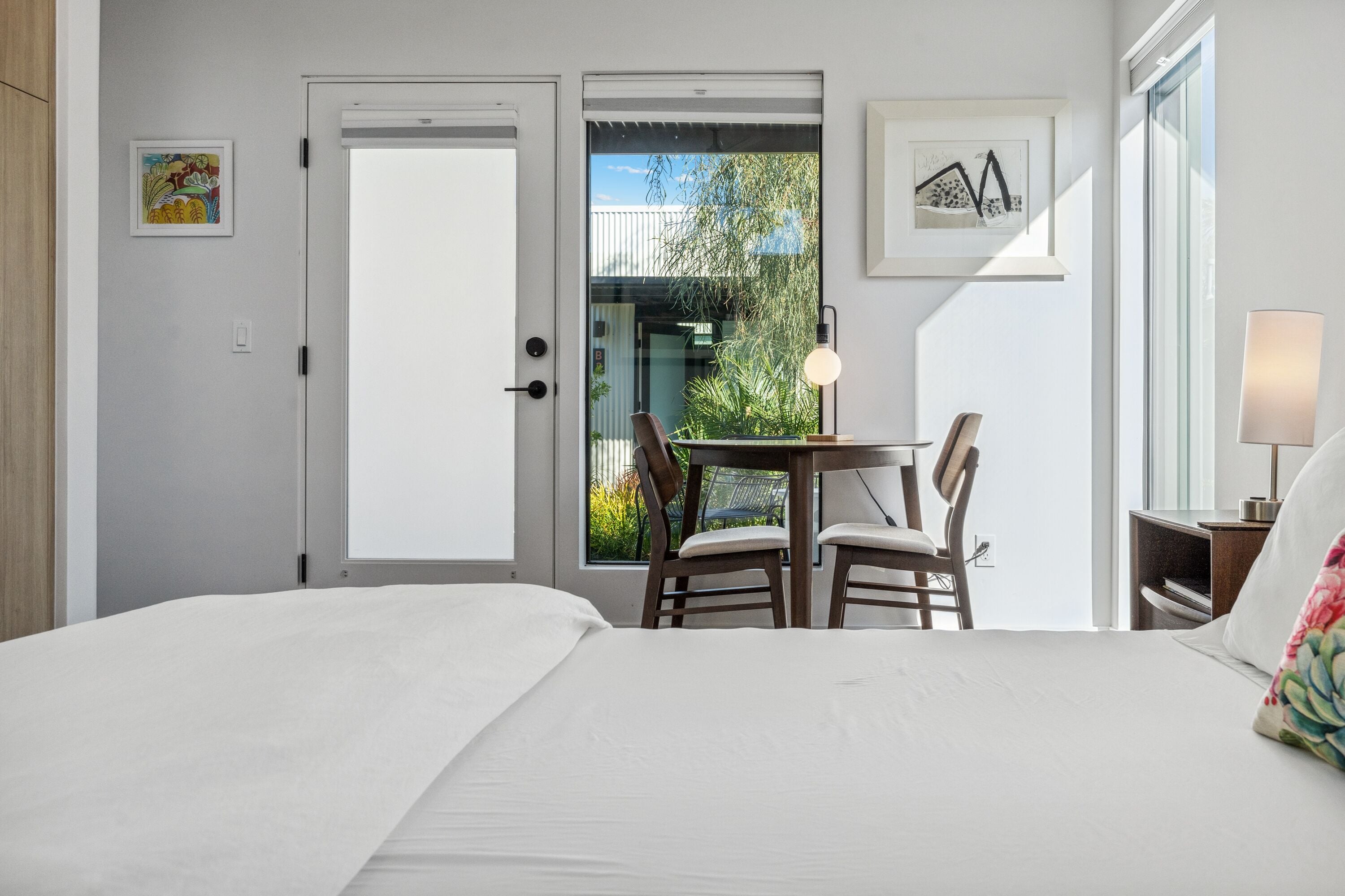 A hotel room with a bed, a table with chairs, a lamp, and a view of the outdoors.