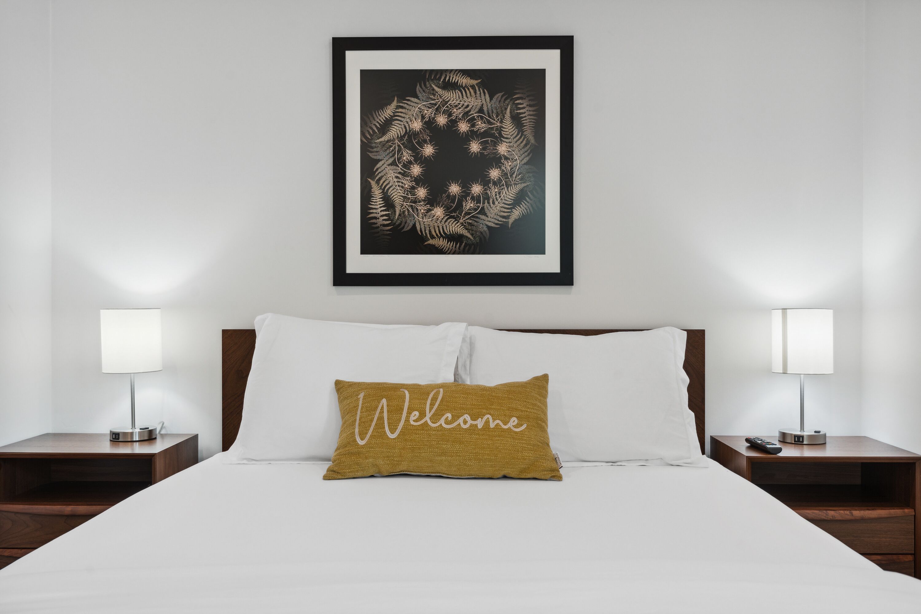 A hotel room with a neatly made bed, a welcome pillow, a framed artwork above the bed, and two bedside tables with lamps.
