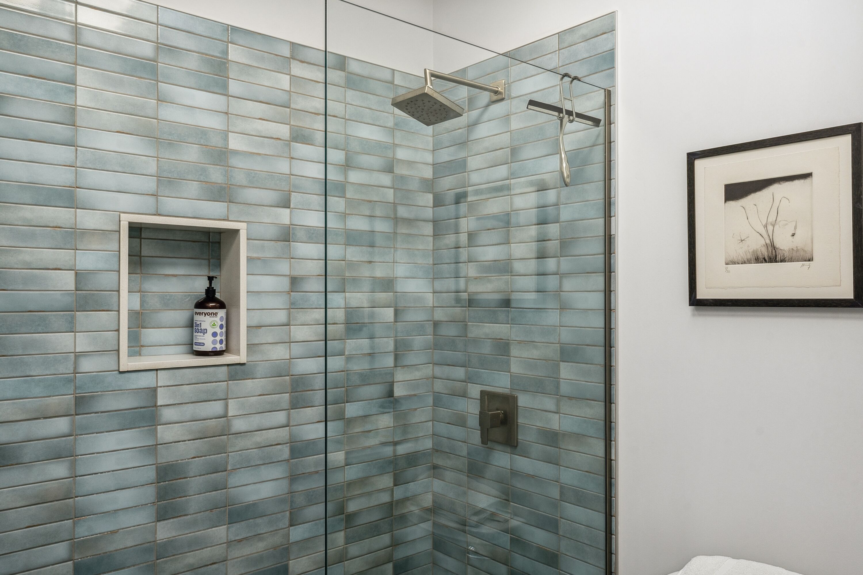 Un baño con pared de ducha en vidrio y mosaicos, un cuadro en la pared y una botella de champú.