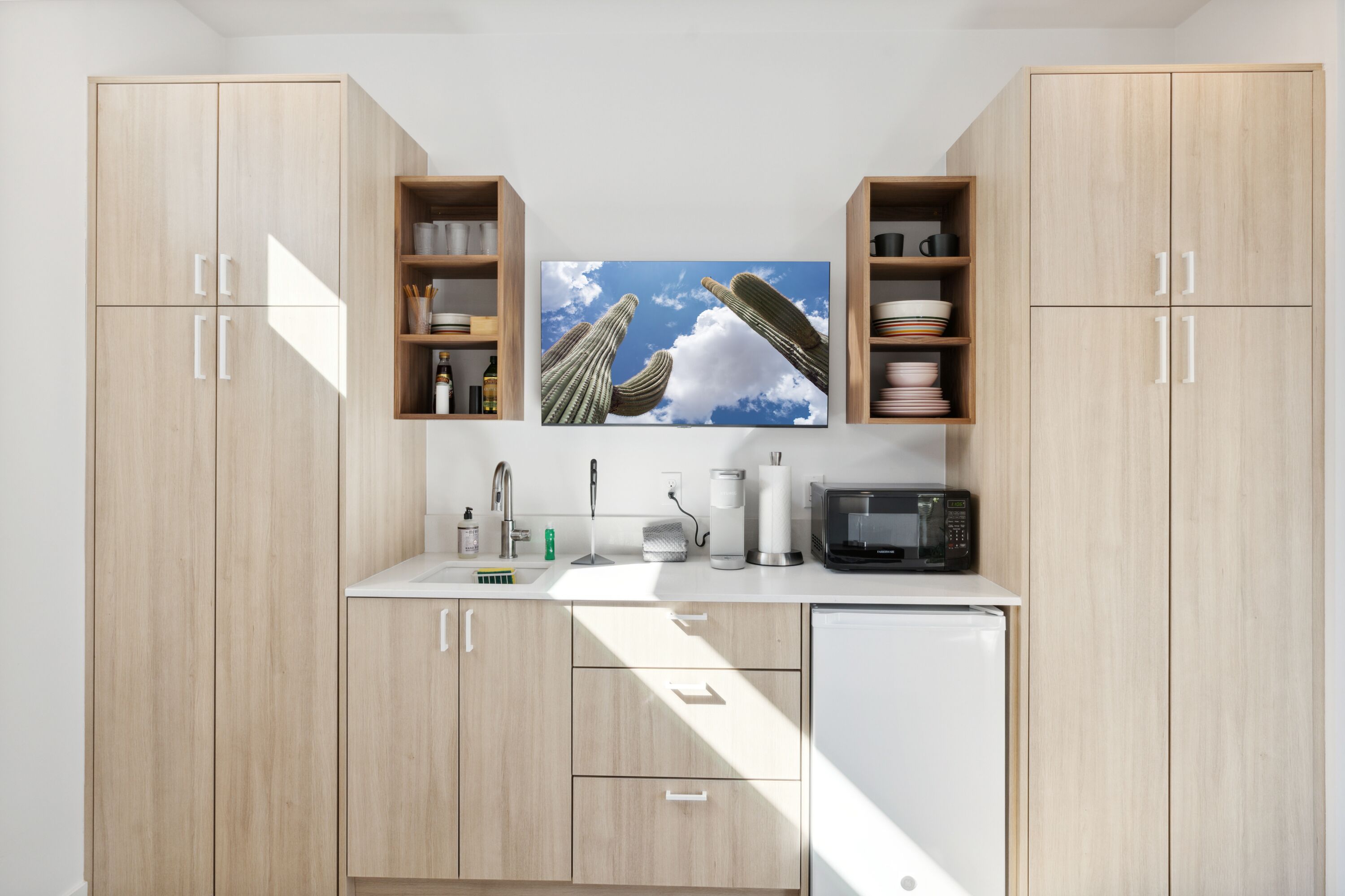 Une cuisine moderne avec des meubles de couleur bois clair, un plan de travail blanc et un grand tableau représentant un cactus au-dessus de l’évier.