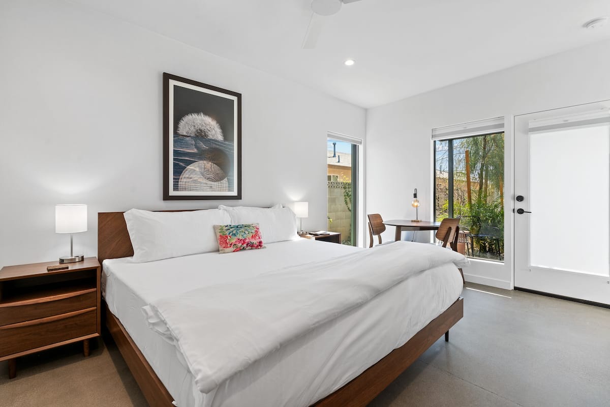 Habitación de hotel moderna con una cama grande, una mesita de noche, un escritorio con una silla y un balcón con vistas a árboles.
