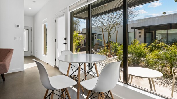 A modern interior with a dining area featuring a round table and four chairs, large windows offering a view of the outdoors, and a minimalist design with white walls and a concrete floor. - 4Square Complex (Phoenix)