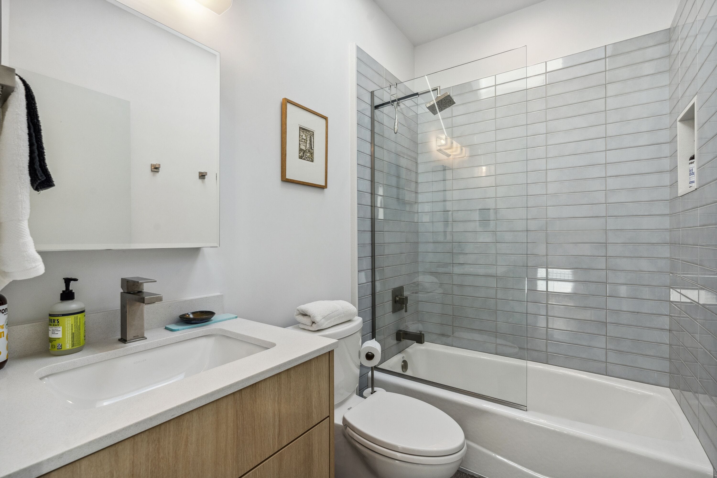 Une salle de bain moderne avec un lavabo, des toilettes et une baignoire, tous de couleur blanche. Un tableau encadré est accroché au mur, et une serviette est suspendue sur le côté gauche.