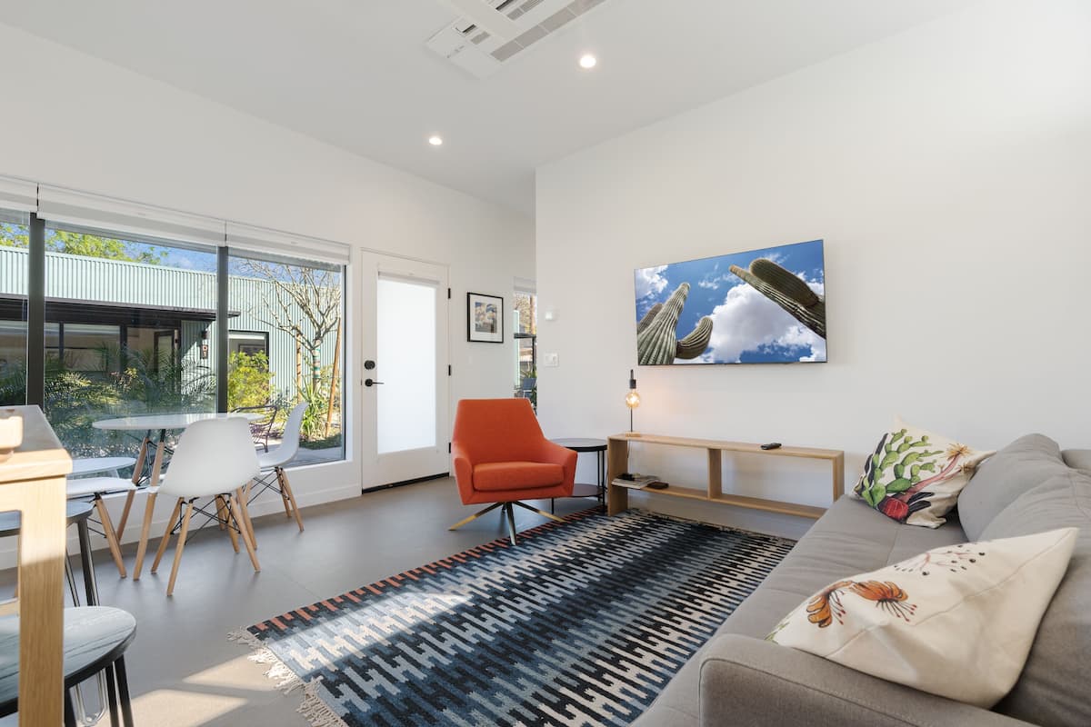 Una sala moderna con un sofá, un sillón rojo y una mesa de centro. Se ve una ventana grande con vistas a un edificio.