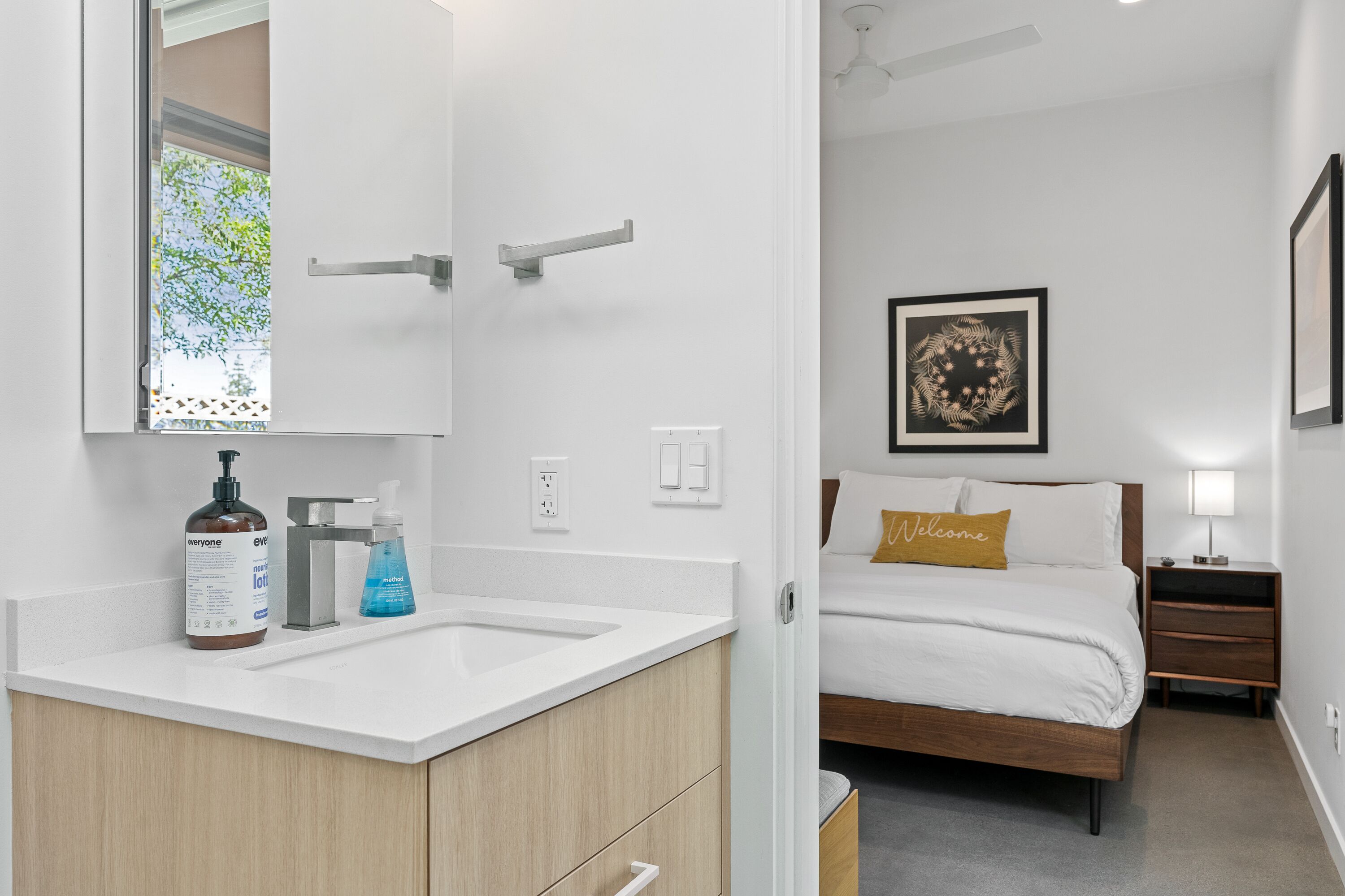 A modern bathroom with a white sink, a mirror, and toiletries. Adjacent is a bedroom with a bed, a nightstand, and a framed artwork on the wall.